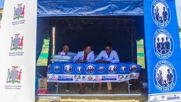 Three people sit under a Republic of Zambia Ministry of Health tent
