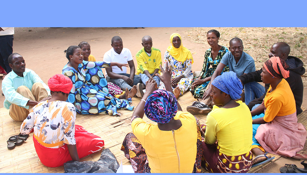 Thirteen members of Juma CHAG sit in a circle on a mat outdoors