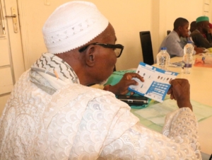 A Nigerian man wearing a white shirt and cap holding a health service referral card.