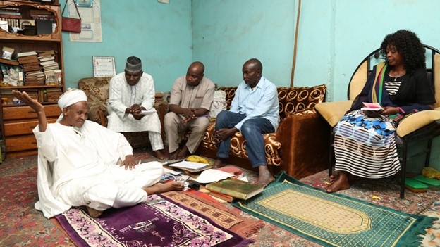 A Nigerian religious leader, sitting on a purple rug, talks to three men and a woman.