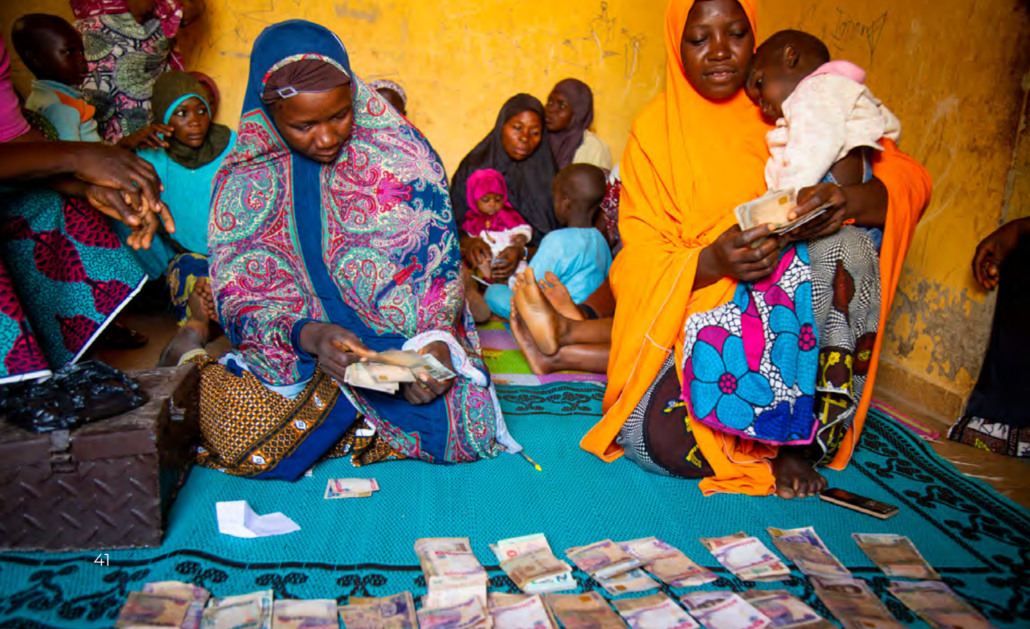 A group of women who are part of a Nigerian women's empowerment group sitting on the ground. The two women in the foreground are counting money.