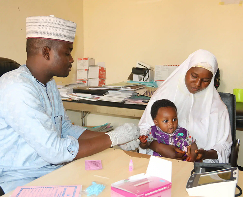 A male health care provider administers a malaria test to a young child sitting on their mother's lap in Nigeria