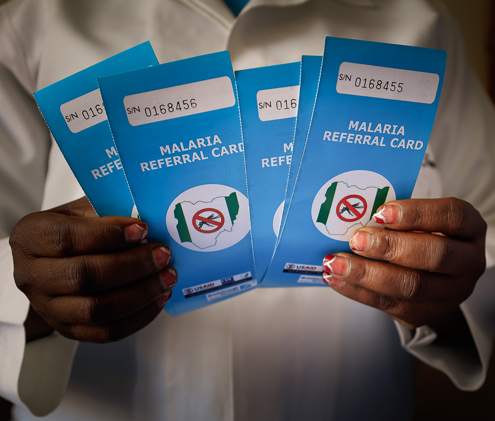 A Nigerian in a white coat holds five malaria referral cards featuring the USAID and Breakthrough ACTION logos.