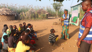 A Nigerian man and woman make a presentation to a group of women and children outside in a village. The woman points to a poster labeled "malaria prevention."