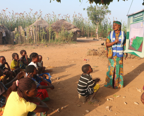 A Nigerian man and woman make a presentation to a group of women and children outside in a village. The woman points to a poster labeled "malaria prevention."