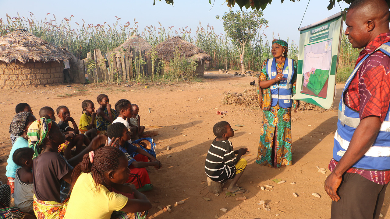 A Nigerian man and woman make a presentation to a group of women and children outside in a village. The woman points to a poster labeled "malaria prevention."