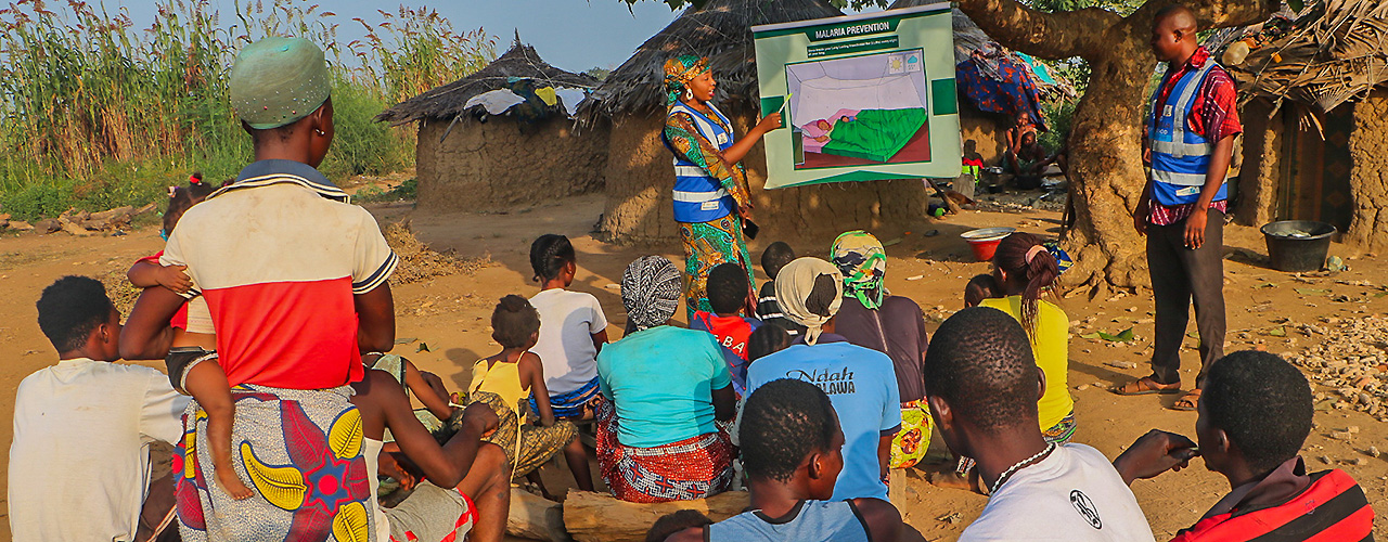 A woman makes a presentation to a group of Nigerian community members, pointing to a poster labeled "malaria prevention." A male presenter looks on.