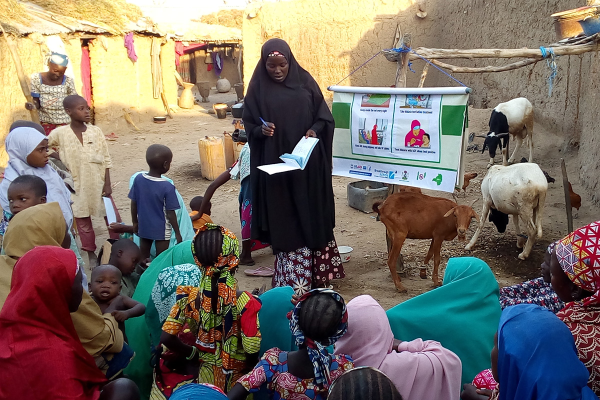 A woman stands, addressing a group of seated mothers and children in a Nigerian village. She holds open a book of referral cards. A flip chart hangs next to the woman, showing Breakthrough ACTION graphics about caring for children with fever.